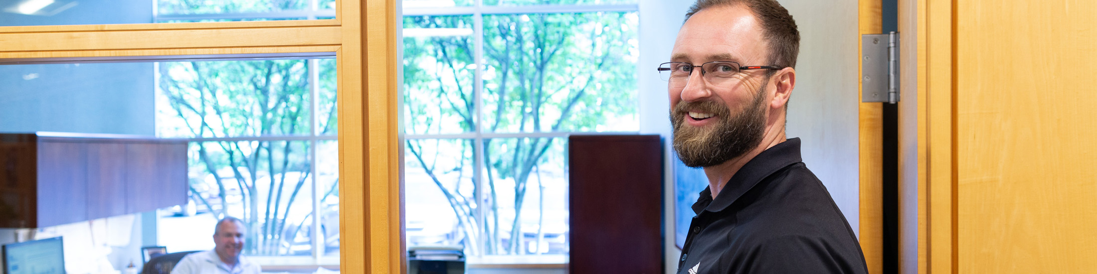 Man looking smiling in doorway of office with man at desk