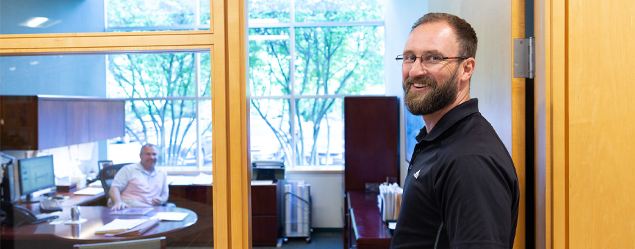 Man looking smiling in doorway of office with man at desk